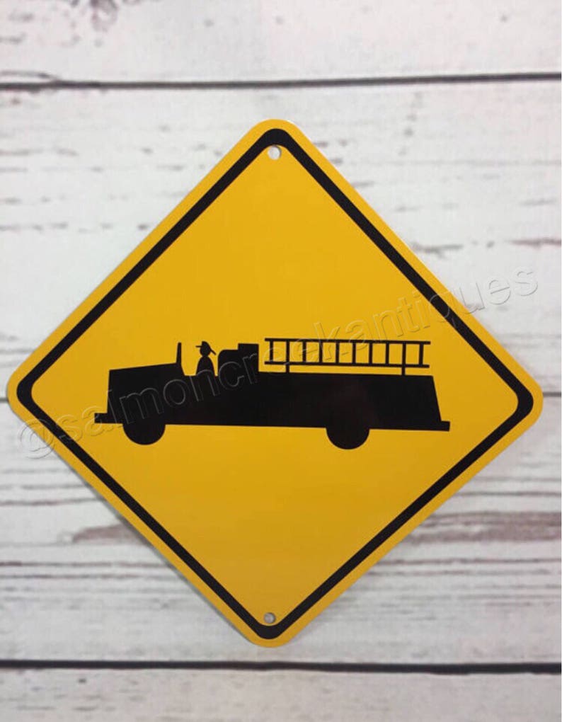 May include: Yellow diamond-shaped road sign with a black border. The sign depicts a black silhouette of a fire truck with a ladder.