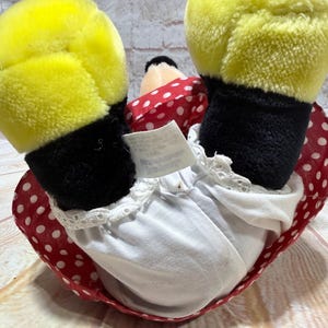 May include: A plush doll featuring yellow gloves, black sleeves, and a red polka-dot dress. The doll's white undergarment has a lace trim. The doll's head is black and white, with a red polka-dot bow.