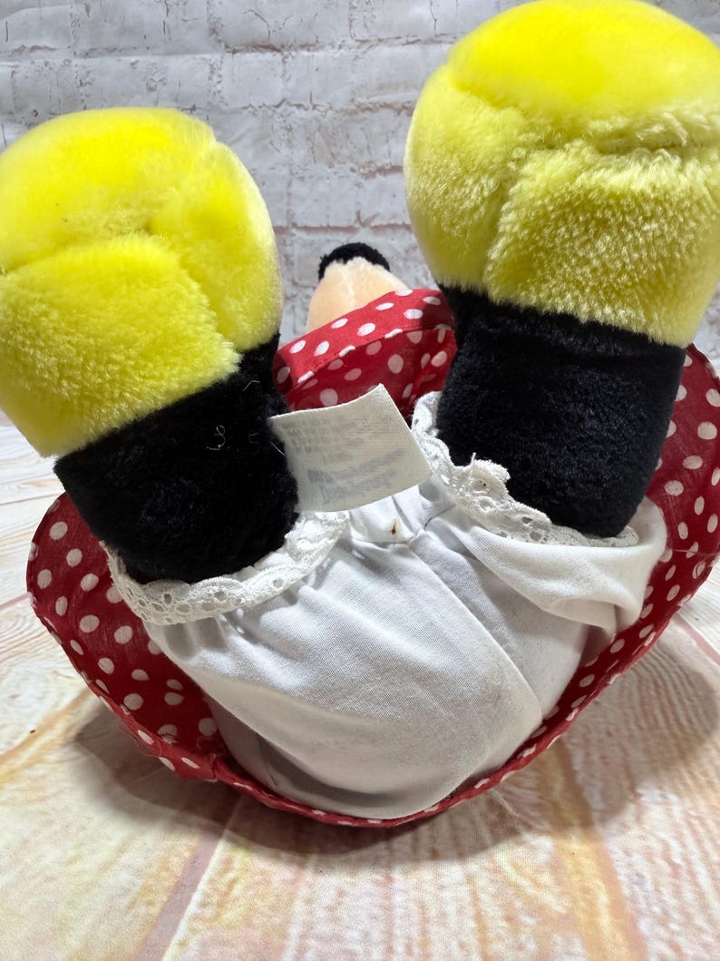 May include: A plush doll featuring yellow gloves, black sleeves, and a red polka-dot dress. The doll's white undergarment has a lace trim. The doll's head is black and white, with a red polka-dot bow.