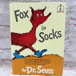 May include: A hardcover children's book titled "Fox in Socks" by Dr. Seuss. The cover features a red fox wearing blue socks, with the title in large black letters. The book is part of the Beginner Books series.