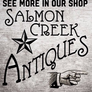 以下が含まれることがあります： 木目調の背景に、スタイリッシュなフォントで「Salmon Creek Antiques」と書かれた白黒の看板。テキストの上に五芒星があり、テキストの下に指さす手が描かれている。