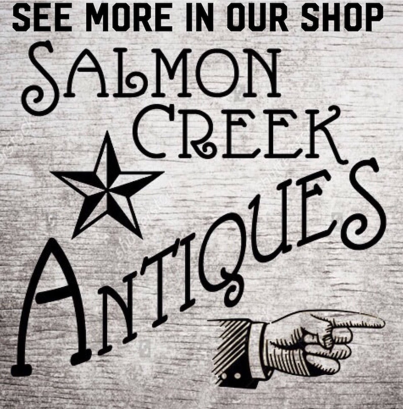 以下が含まれることがあります： 木目調の背景に、スタイリッシュなフォントで「Salmon Creek Antiques」と書かれた白黒の看板。テキストの上に五芒星があり、テキストの下に指さす手が描かれている。