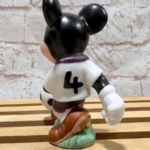 Pu&ograve; includere: Statuina di un topo dei cartoni animati in divisa da calcio. Il topo indossa una maglia bianca con il numero 4, pantaloncini neri e scarpe marroni. Tiene in mano un pallone da calcio e si trova su un prato verde.