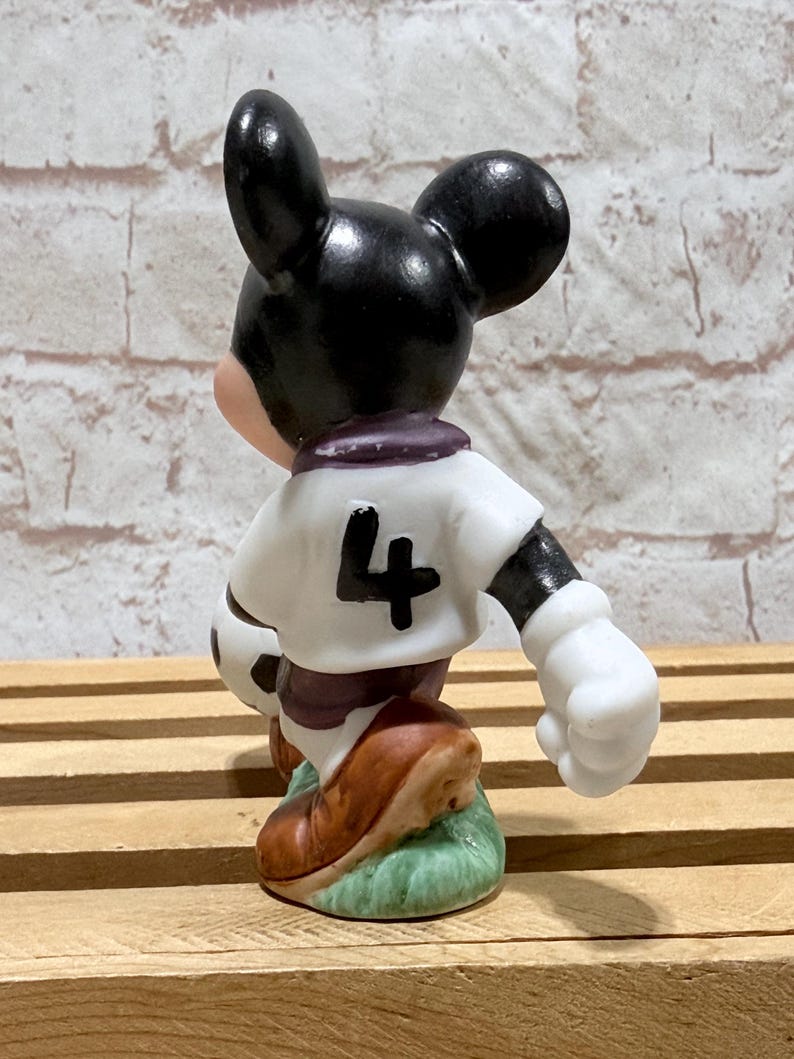 Pu&ograve; includere: Statuina di un topo dei cartoni animati in divisa da calcio. Il topo indossa una maglia bianca con il numero 4, pantaloncini neri e scarpe marroni. Tiene in mano un pallone da calcio e si trova su un prato verde.