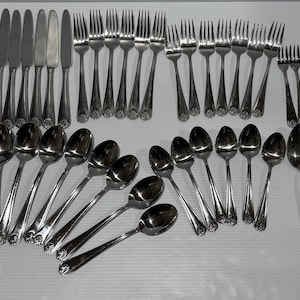 May include: A collection of stainless steel silverware, including knives, forks, and spoons, arranged on a white surface. The set includes various spoon sizes, butter knives, and forks, ideal for a formal dining setting. The silverware has a decorative floral design.