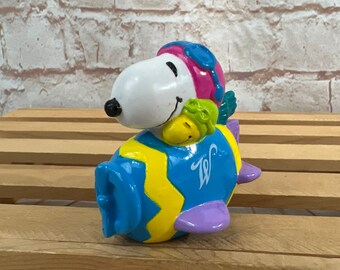 Può includere: Una statuetta in ceramica di Snoopy, il personaggio dei Peanuts, che pilota un aereo blu e giallo con una coda viola. Snoopy indossa un casco da pilota rosa e blu e occhiali protettivi. L'aereo ha una 'W' gialla sul lato.