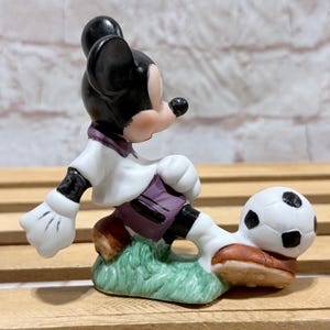 Pu&ograve; includere: Statuetta in ceramica di Topolino che gioca a calcio. Topolino indossa una camicia bianca con dettagli viola, pantaloncini neri e scarpe marroni. Il pallone da calcio &egrave; bianco e nero e la statuetta &egrave; su una base verde.