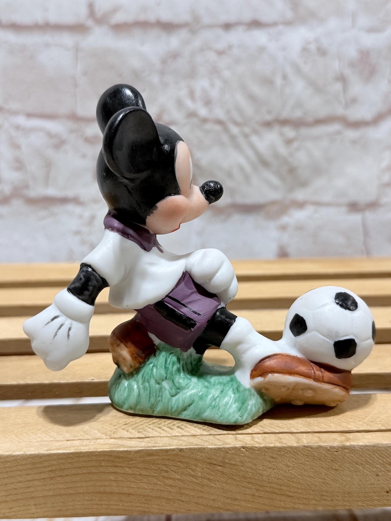 Pu&ograve; includere: Statuetta in ceramica di Topolino che gioca a calcio. Topolino indossa una camicia bianca con dettagli viola, pantaloncini neri e scarpe marroni. Il pallone da calcio &egrave; bianco e nero e la statuetta &egrave; su una base verde.