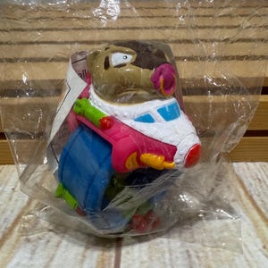 May include: A colorful plastic toy figure of a cartoon character, packaged in a clear plastic bag. The figure has a brown head, pink nose, and is wearing a blue, white, and red outfit. The toy is likely a collectible or part of a children's play set.