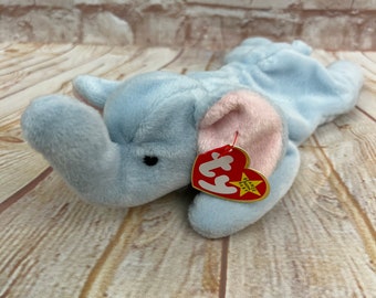 May include: A light blue plush elephant toy with pink inner ears and a red heart-shaped tag. The elephant is lying down on a wooden surface, with a brick wall in the background. The tag reads "Beanie Baby".