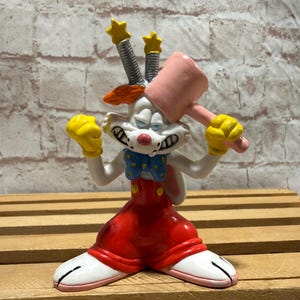 May include: A figurine of a cartoon rabbit with a pink mallet, yellow gloves, and red overalls. The rabbit has a blue bow tie and is holding the mallet above its head. The figurine has a white face and ears, and is standing on a wooden surface.