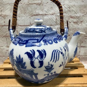May include: A white ceramic teapot with a blue willow pattern. The teapot has a woven brown handle and a matching lid. The spout and body are decorated with detailed blue illustrations. The teapot is sitting on a wooden surface.