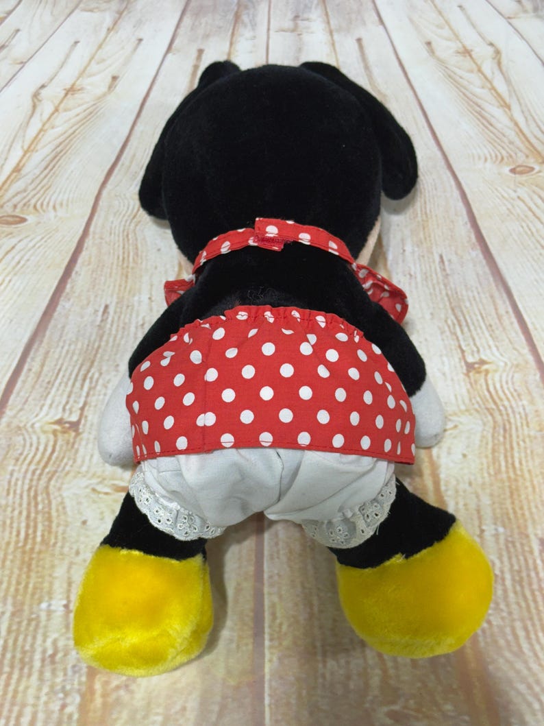 K&ouml;nnte beinhalten: Pl&uuml;schtier einer Zeichentrickfigur, von hinten gesehen. Die Figur hat einen schwarzen Kopf und Ohren, einen roten Polka-Dot-Rock und wei&szlig;e Bloomers mit Spitzenbesatz. Gelbe F&uuml;&szlig;e und eine rote Polka-Dot-Schleife vervollst&auml;ndigen den Look.