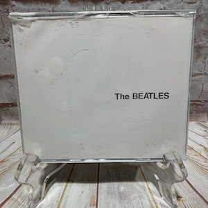 May include: A CD case with a white cover and the band name "The BEATLES" printed in black. The case is displayed on a clear plastic stand against a backdrop of a brick wall and wooden planks.