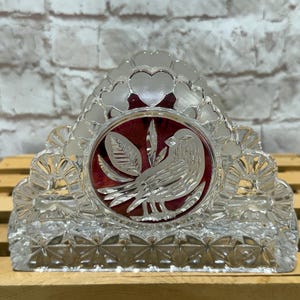 May include: A clear glass napkin holder featuring a central circular medallion with a deep red background and a silver-toned etched bird design. The frame is decorated with a repeating heart pattern along the top edge and a geometric star motif.