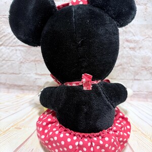 May include: A plush Minnie Mouse doll, viewed from the back, features a black body, large round ears, and a red and white polka dot skirt. A red bow with white polka dots adorns the back of the neck. The doll is a classic children's toy.