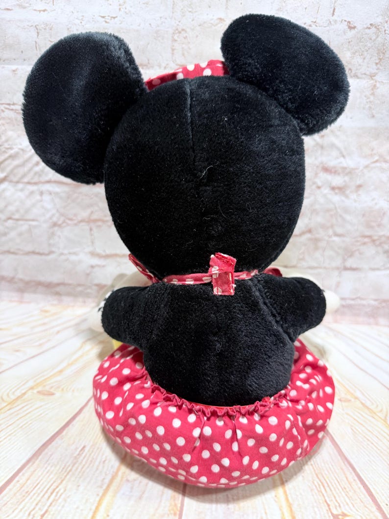 May include: A plush Minnie Mouse doll, viewed from the back, features a black body, large round ears, and a red and white polka dot skirt. A red bow with white polka dots adorns the back of the neck. The doll is a classic children's toy.