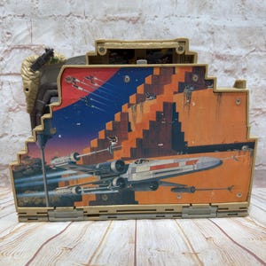 May include: A vintage Star Wars action figure playset featuring a detailed illustration of X-wing fighters in flight against a sunset backdrop. The playset is tan and features multiple compartments and hinged panels.