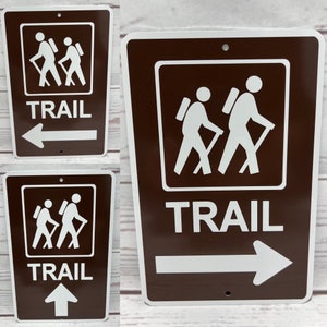 May include: Three brown metal signs with white text and graphics. Each sign says "TRAIL" and has a graphic of two hikers with backpacks. The signs have arrows pointing in different directions: left, right, and up.