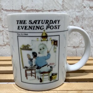 May include: White ceramic mug featuring a vintage illustration from The Saturday Evening Post, dated Feb. 13, 1960. The artwork depicts an artist at work. The mug has a curved handle and is set against a light-colored background.