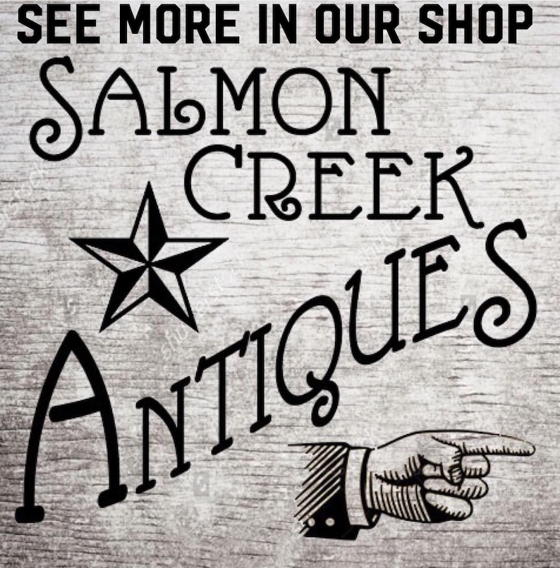 Pu&ograve; includere: Un cartello in legno con la scritta "SEE MORE IN OUR SHOP" sopra "SALMON CREEK ANTIQUES". Sono inclusi anche una stella nera e una grafica di una mano che indica. Il cartello ha un aspetto rustico e invecchiato.