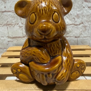 Può includere: Vaso di miele a forma di orso in ceramica marrone. L'orso è seduto e tiene in mano un vaso di miele. Il vaso di miele è di colore marrone lucido. L'orso ha grandi occhi rotondi e piccole orecchie. Il vaso di miele è su un pallet di legno.
