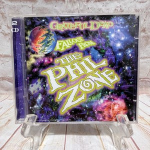 Può includere: Un CD musicale dei Grateful Dead intitolato "Fallout From The Phil Zone". La copertina dell'album presenta un colorato design cosmico con il nome della band e il titolo dell'album in lettere vivaci. La custodia del CD è esposta su un supporto di plastica trasparente.