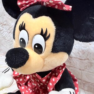 May include: A plush Minnie Mouse doll with a black head, ears, and body. The doll has a cream-colored face, a black nose, and large eyes. She wears a red dress and bow, both with white polka dots. The doll's hands are white.