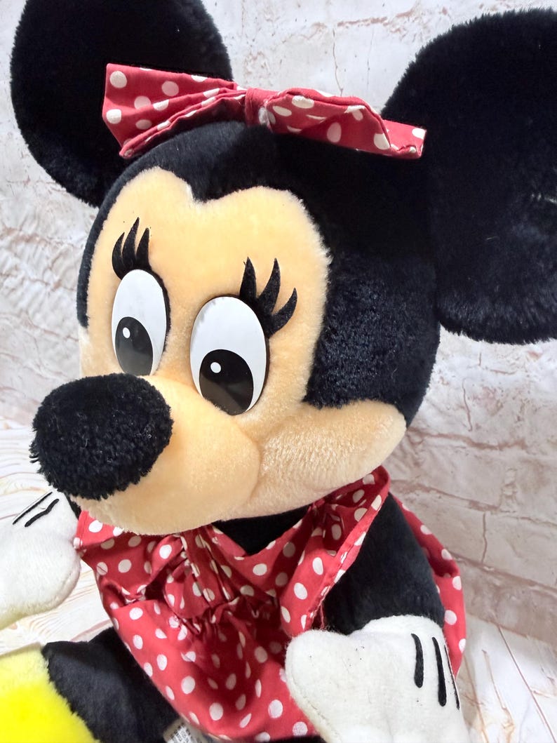 May include: A plush Minnie Mouse doll with a black head, ears, and body. The doll has a cream-colored face, a black nose, and large eyes. She wears a red dress and bow, both with white polka dots. The doll's hands are white.