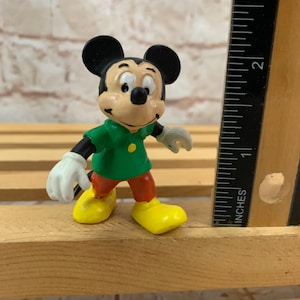 以下が含まれることがあります： 緑のシャツと赤いズボンを着たミッキーマウスの小さなプラスチック製フィギュア。彼は、背景に定規のある木製の表面に立っています。