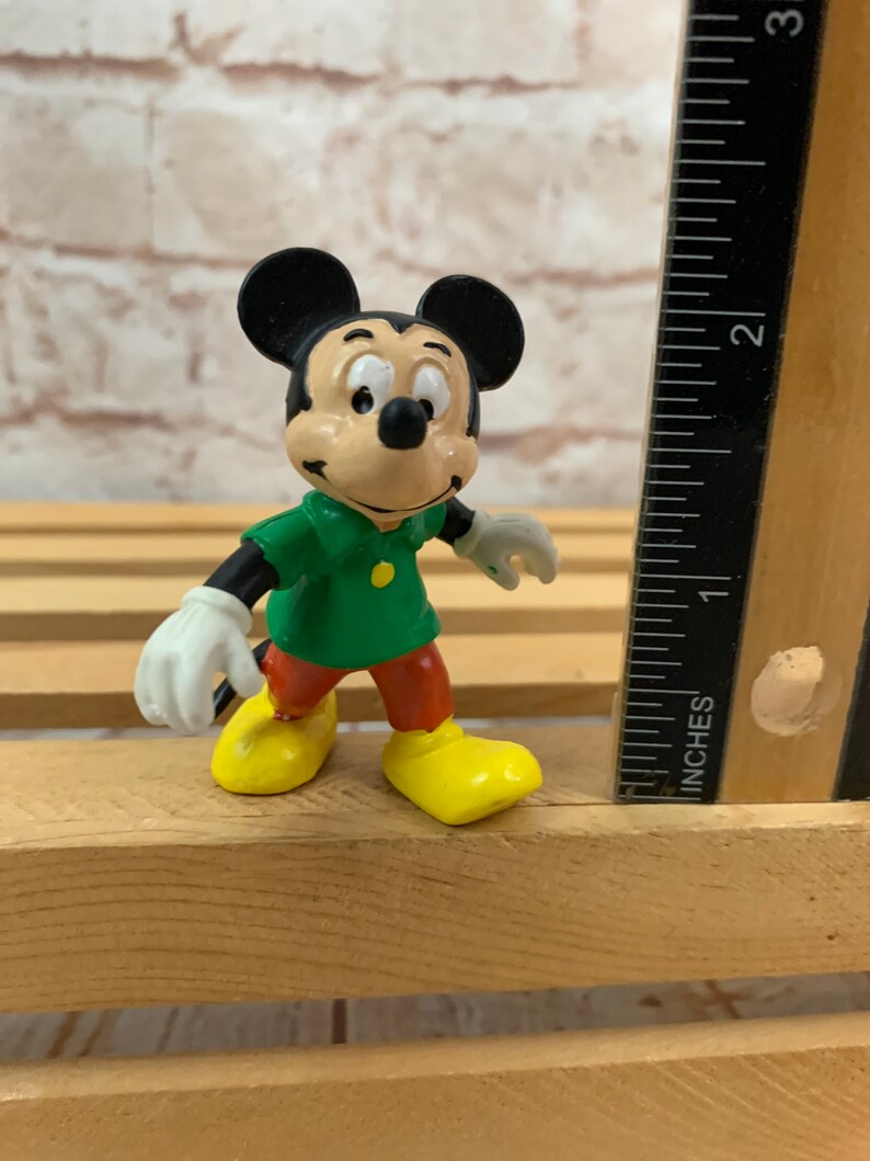 以下が含まれることがあります： 緑のシャツと赤いズボンを着たミッキーマウスの小さなプラスチック製フィギュア。彼は、背景に定規のある木製の表面に立っています。