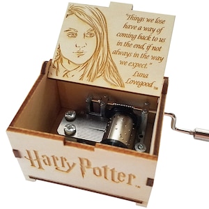 May include: A wooden music box with a hand-crank on the side. The lid is open and features a laser-engraved portrait of Luna Lovegood from the Harry Potter series. The inscription reads "Things we lose have a way of coming back to us in the end, if not always in the way we expect." Luna Lovegood TM. The bottom of the box is engraved with "Harry Potter TM."