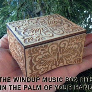 K&ouml;nnte beinhalten: Eine kleine Holz-Musikbox mit einem komplizierten floralen Design, das in den Deckel und die Seiten geschnitzt ist. Die Box ist klein genug, um in die Handfl&auml;che zu passen. "THE WINDUP MUSIC BOX FITS IN THE PALM OF YOUR HAND" steht unter der Box.