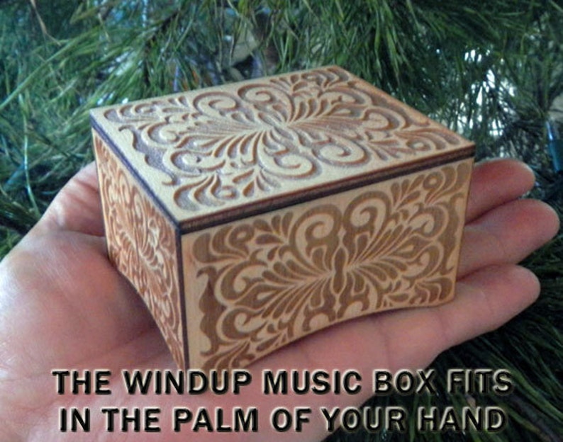 K&ouml;nnte beinhalten: Eine kleine Holz-Musikbox mit einem komplizierten floralen Design, das in den Deckel und die Seiten geschnitzt ist. Die Box ist klein genug, um in die Handfl&auml;che zu passen. "THE WINDUP MUSIC BOX FITS IN THE PALM OF YOUR HAND" steht unter der Box.