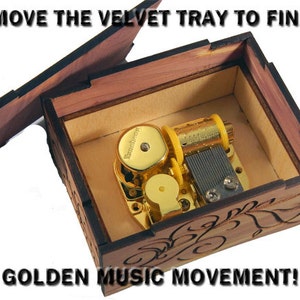 May include: A wooden music box with a hinged lid open, revealing a gold-colored music movement inside. The box has a carved floral design and the text "REMOVE THE VELVET TRAY TO FIND: GOLDEN MUSIC MOVEMENT!" is printed on the lid.
