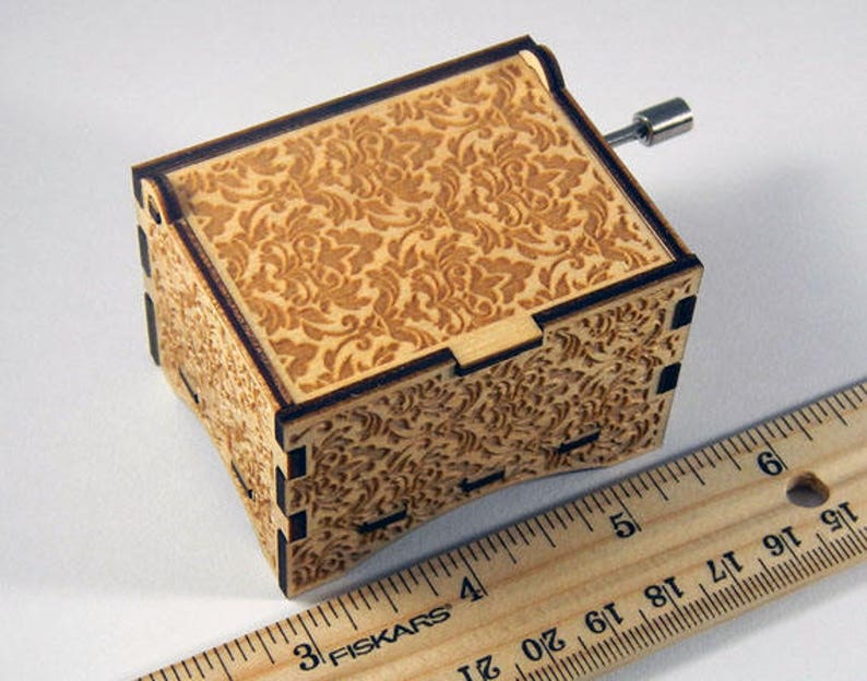 May include: A wooden music box with a floral pattern on the top and sides. The box is closed and has a metal winding key on the side.