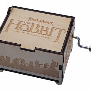 May include: Wooden music box featuring the text "The Hobbit" and "Lord of the Rings" with a crank on the side. The box has a laser-cut design of characters from the movie. The box is made of light-colored wood.