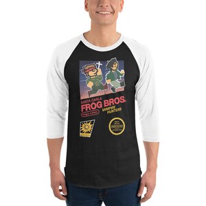 Puede incluir: Camiseta de béisbol raglán en blanco y negro con un gráfico de la película "The Lost Boys". El gráfico presenta personajes pixelados y el texto "Santa Carla Frog Bros. Vampire Hunters."
