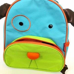 May include: A blue and green Skip Hop Zoo Pack backpack shaped like a dog with brown ears and a black nose. The backpack has a zippered opening and a small pocket on the front.