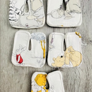 Può includere: Cinque lettere in tessuto bianco che compongono la parola "WOOHOO" con i personaggi di Winnie the Pooh e i suoi amici su ogni lettera. Le lettere sono cucite insieme con un filo bianco.