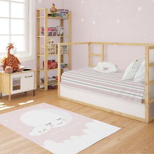 Può includere: Un tappeto per bambini rosa e bianco con un design a forma di luna e nuvole. Il tappeto si trova in una camera da letto con un letto in legno, un peluche a forma di giraffa e uno scaffale in legno con giocattoli.