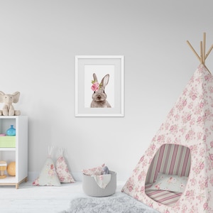 May include: A white wall with a white frame picture of a rabbit with a pink flower crown. There is a white shelf with a stuffed elephant, a blue vase, and a green box. A pink and white floral teepee with a striped mattress is in the foreground.