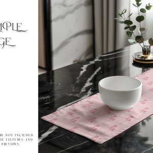 May include: A pink table runner with a delicate floral pattern sits on a black marble table. A white bowl is placed on the runner, and a small potted plant is in the background.