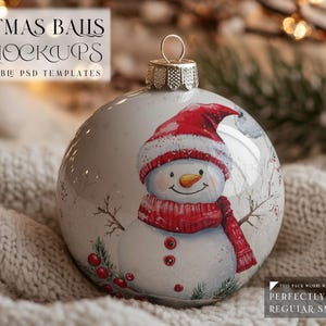 May include: A white Christmas ornament with a snowman design. The snowman is wearing a red hat and scarf. The ornament is sitting on a white knitted blanket.