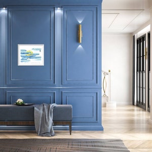 May include: A blue wall with white trim and gold wall lights. A framed abstract painting with blue and yellow paint is hanging on the wall. A bench with a grey blanket is in front of the wall.