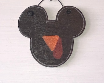 May include: A wooden, dark brown Mickey Mouse-shaped ornament with a light gray outline. The ornament features a triangle-shaped nose and tongue in shades of orange and brown. A small metal loop is attached for hanging.