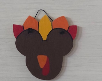 May include: A wooden Thanksgiving turkey decoration with a brown body and colorful feathers in shades of red, orange, and yellow. The turkey has a black string for hanging.