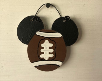 May include: A decorative wooden football-shaped ornament with black circular ears. The football is brown with white stripes and a white design in the center. The ornament hangs from a black wire loop.