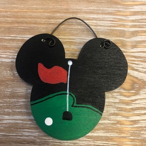 May include: A black wooden golf-themed decoration in the shape of Mickey Mouse's head. The design features a green golf course, a red flag, and a white golf ball. A black wire loop is attached for hanging.