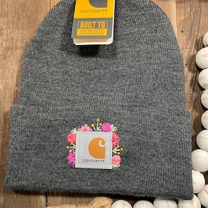 May include: A dark gray knit beanie with a folded cuff featuring a square Carhartt patch embroidered with colorful pink, yellow, and green floral accents. A yellow and black Carhartt brand tag is attached to the top of the beanie.